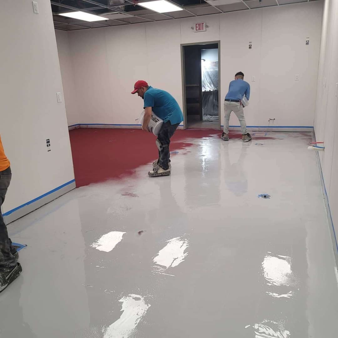 Workers applying epoxy floor coating in a commercial space with red and gray paint.