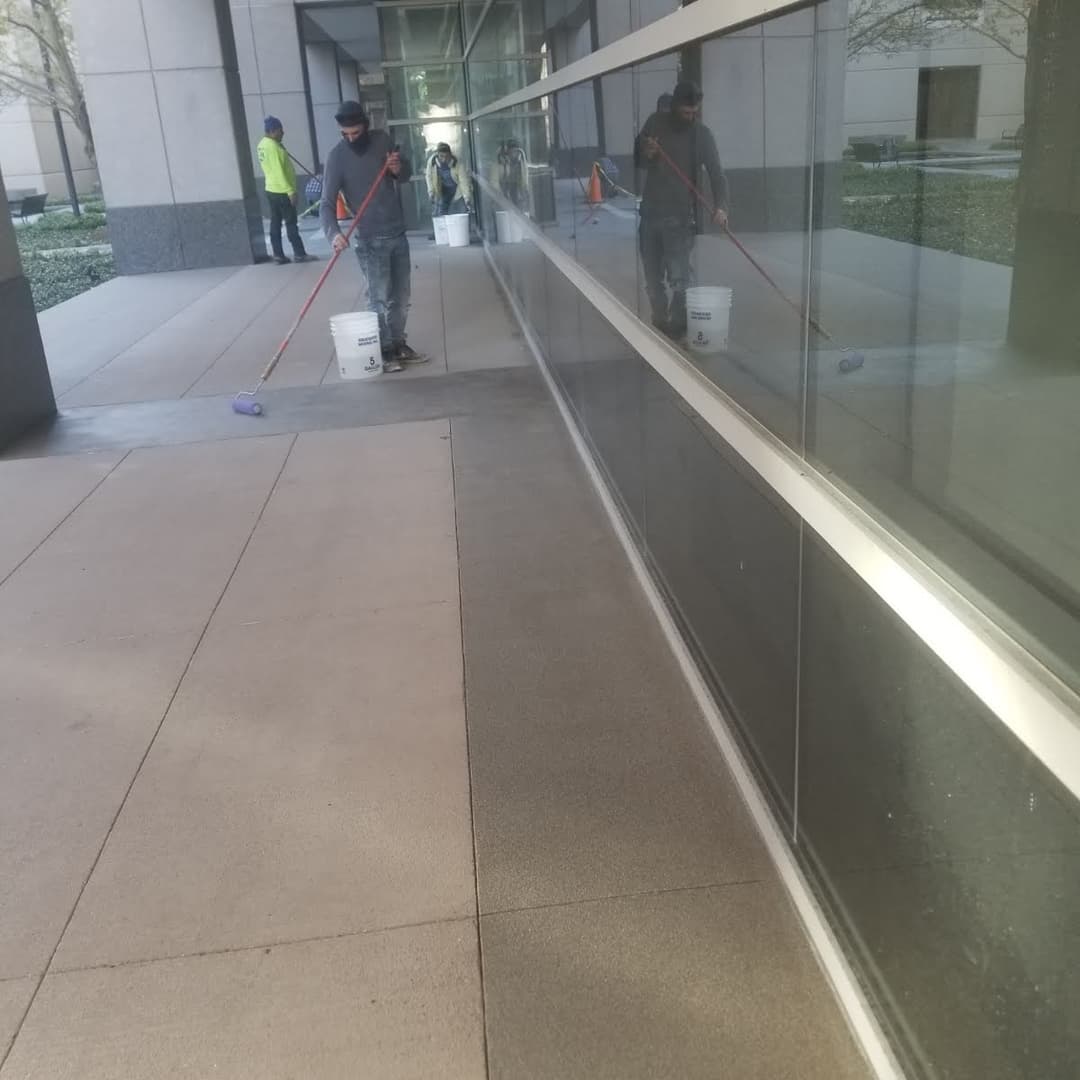 Worker painting a sidewalk with a roller next to glass building reflecting surroundings.