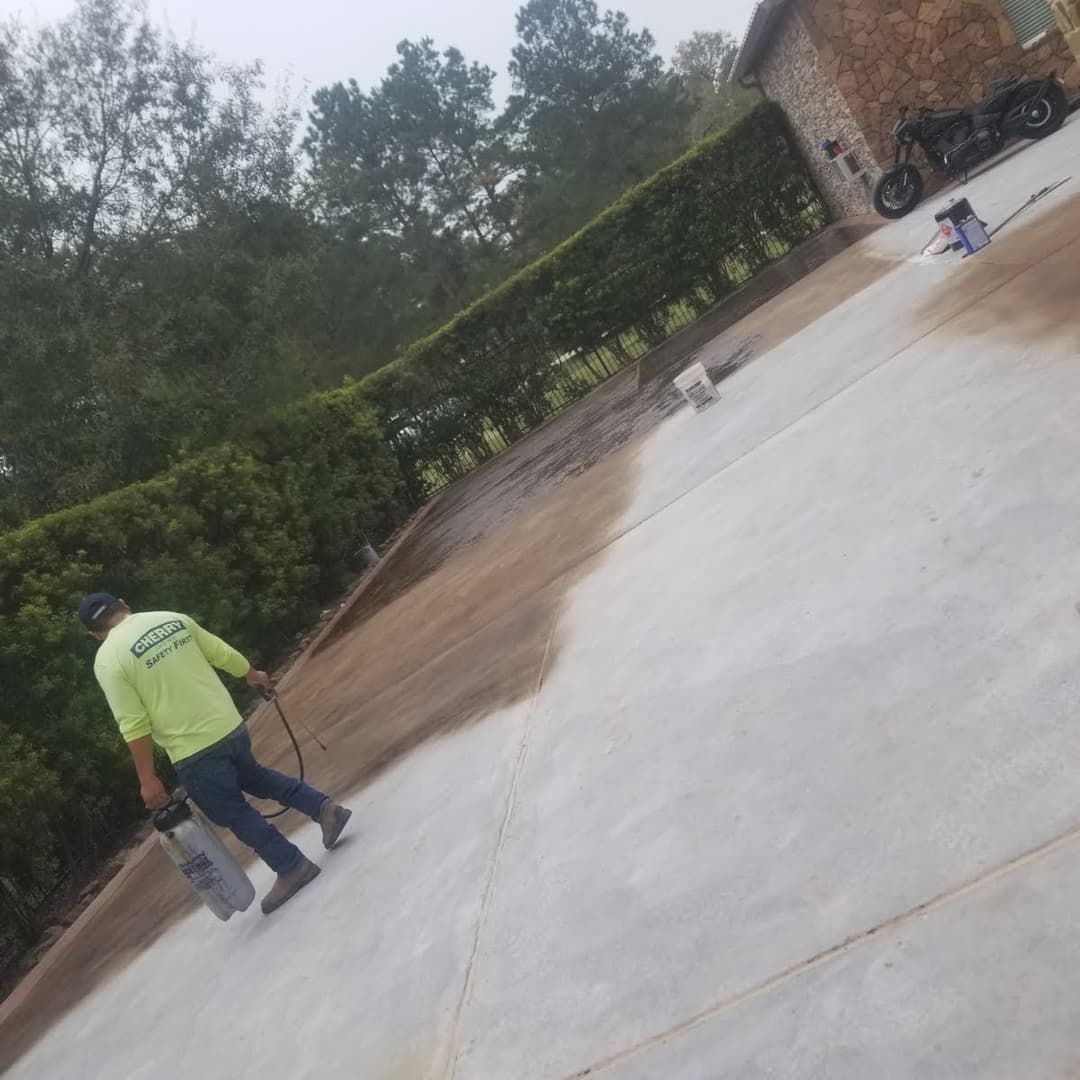 Worker pressure washing a concrete driveway beside a motorcycle and greenery.