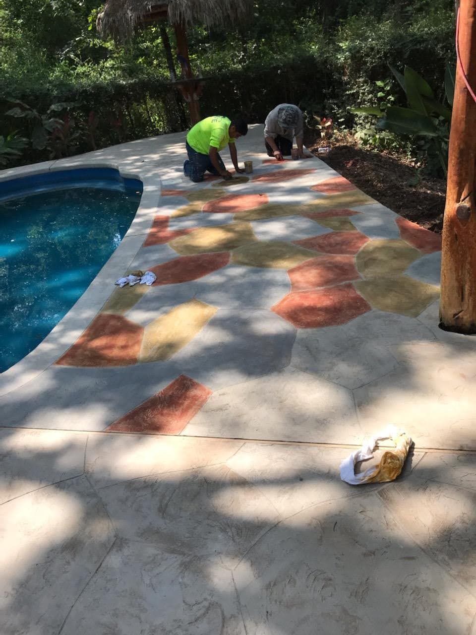 Workers applying decorative concrete near a pool in a lush outdoor setting.