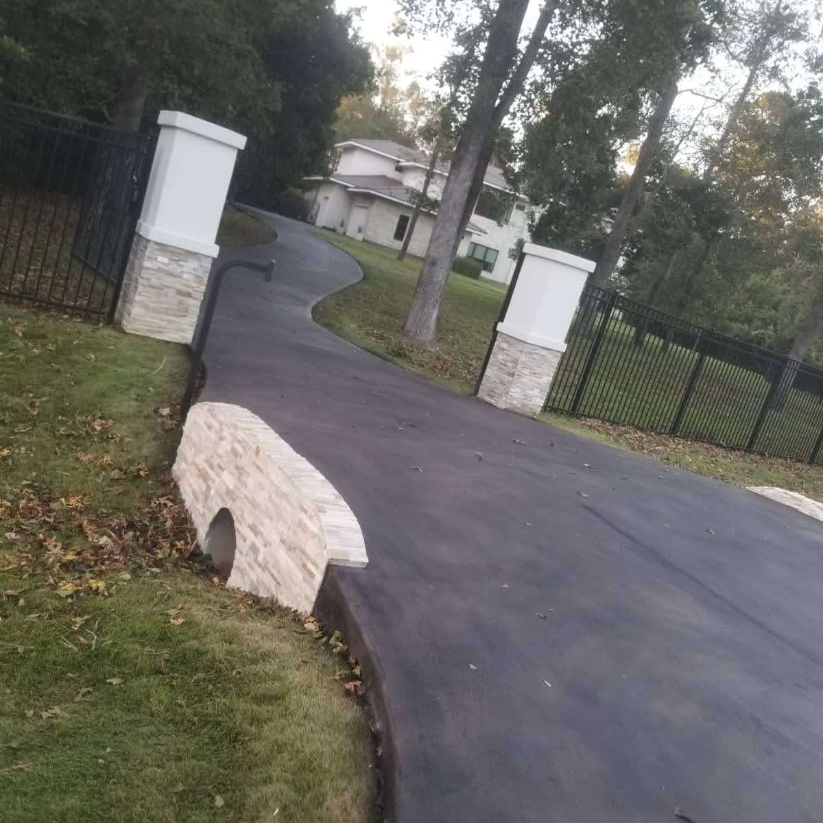Curved driveway entrance with stone pillars and a modern house in a wooded area.