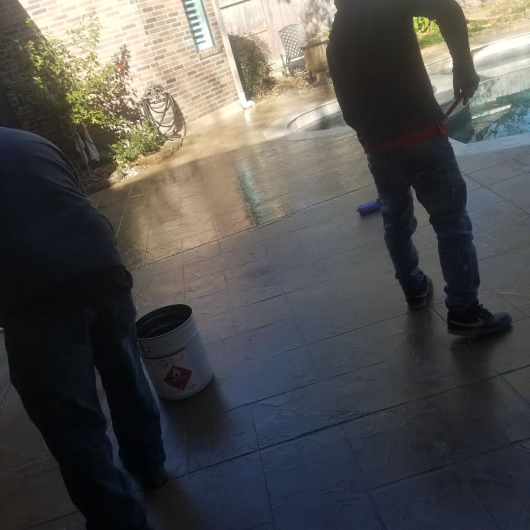 Workers applying sealant on a stamped concrete patio near a pool.