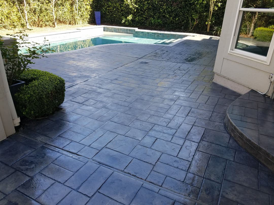 Stylish stamped concrete patio beside a pool, surrounded by lush greenery and shrubs.