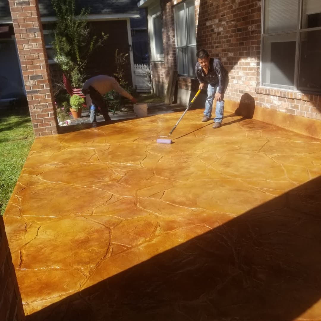 Workers applying decorative concrete sealant on a textured patio in a residential backyard.