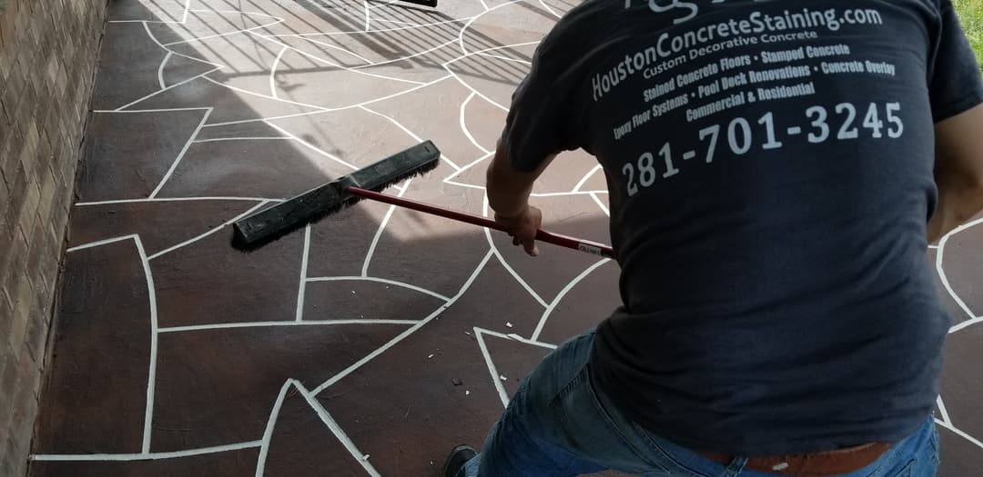 Person applying decorative concrete stain on outdoor surface with a long-handled tool.