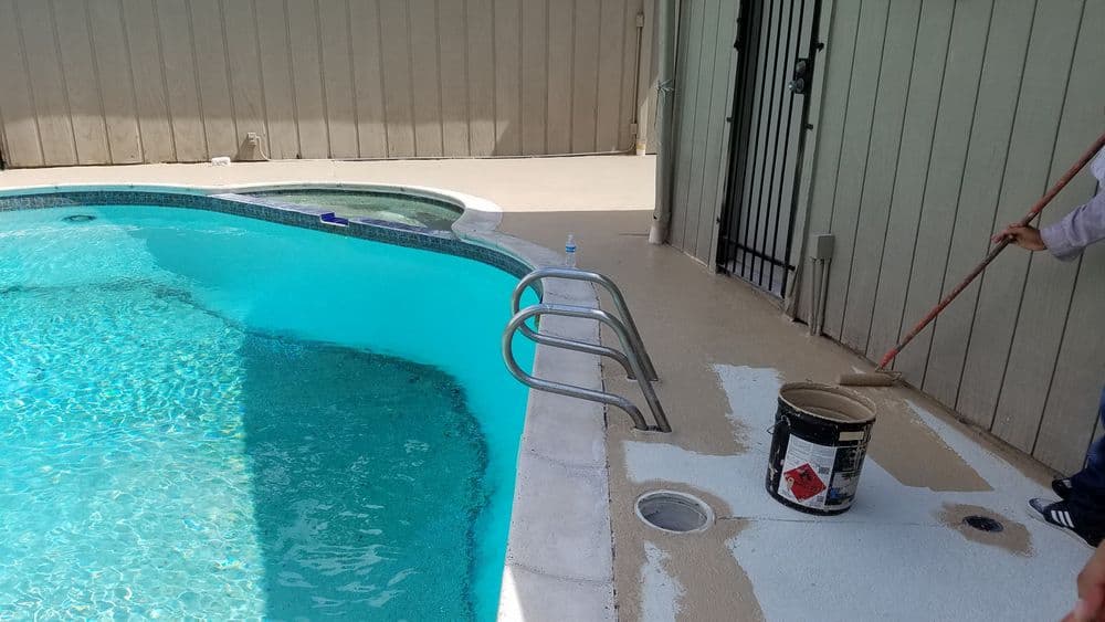 Person painting the pool deck beside a clear blue pool with a roller and paint can.