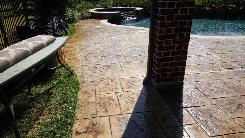 Decorative stamped concrete patio surrounding a pool with landscaping in sunny backyard setting.