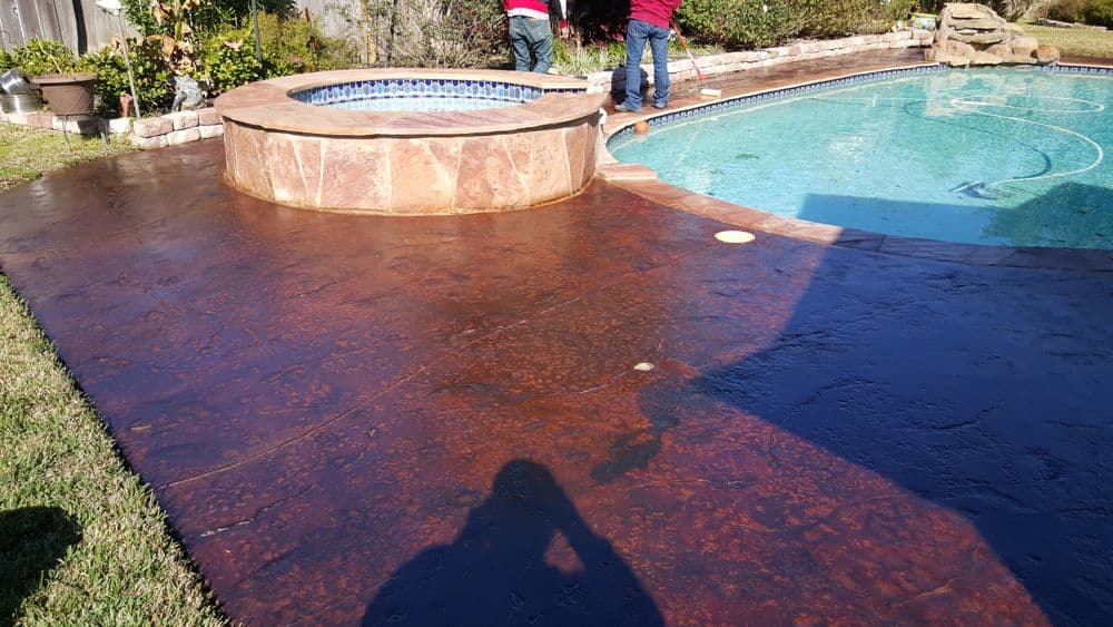 Workers applying a sealant on a decorative concrete patio around a pool and spa.